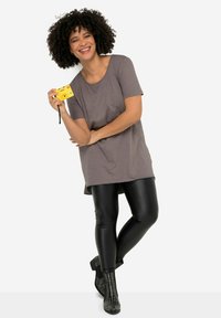 T-shirt gris oversize, leggings noirs brillants et bottines noires à clous. Appareil photo jaune tenu d'une main, pose détendue.