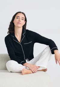 Chemise de pyjama noire à boutons avec des détails en piping blancs, associée à un pantalon blanc texturé ample. Modèle assis sur une surface douce.