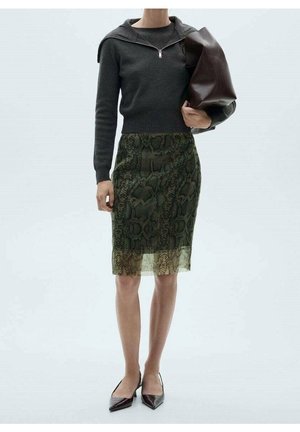 Femme portant un haut à manches longues gris foncé, une jupe verte à imprimé serpent longueur genoux, des chaussures pointues noires, tenant un grand sac à main marron foncé.