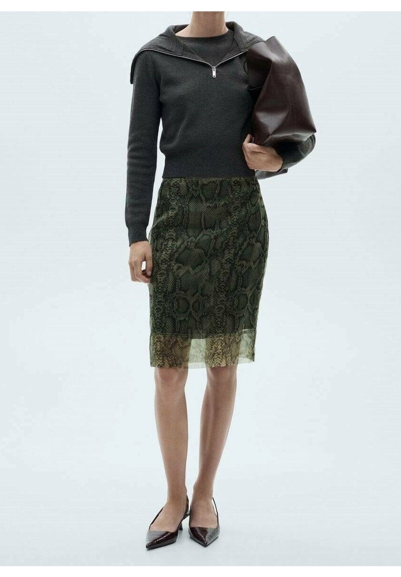 Femme portant un haut à manches longues gris foncé, une jupe verte à imprimé serpent longueur genoux, des chaussures pointues noires, tenant un grand sac à main marron foncé.