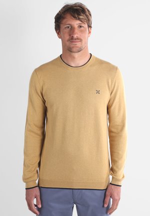 Pull en tricot jaune moutarde avec manches longues, col rond et ourlet côtelé. Petit logo foncé sur la poitrine gauche, texture lisse.