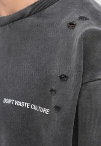 Sudadera gris oscuro con pequeños agujeros cerca del hombro y texto blanco que dice "NO DESPERDICIES CULTURA" en el pecho.