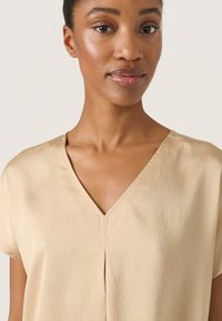 Beige V-ringad blus med korta ärmar, med ett subtilt strukturerat mönster och en centralt placerad sömdetalj. Mjukt tyg med en slät yta.