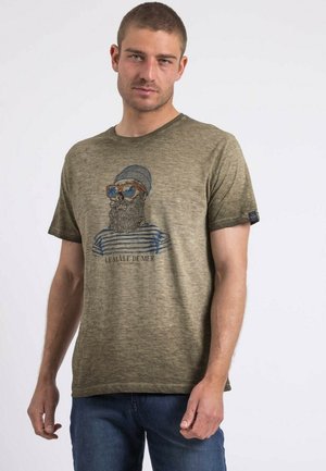 T-shirt vert olive avec un graphique d'un homme barbu portant des lunettes de soleil, avec un motif rayé bleu. Tissu doux, col rond, manches courtes.