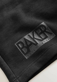 Zbliżenie na czarną tkaninę z przeszytym obrębem i błyszczącą prostokątną naszywką z napisem "BAKER" drukowanymi, dużymi literami.