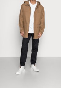 Bruine wollen mix jas met een capuchon, drukknoopsluitingen en twee voorzakken, gecombineerd met zwarte broeken en witte sneakers op een lichte achtergrond.