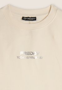 Sweatshirt beige avec col côtelé, orné d'un texte imprimé argenté : "FREDDY AUJOURD'HUI EST TON JOUR." Texture douce, coupe décontractée.