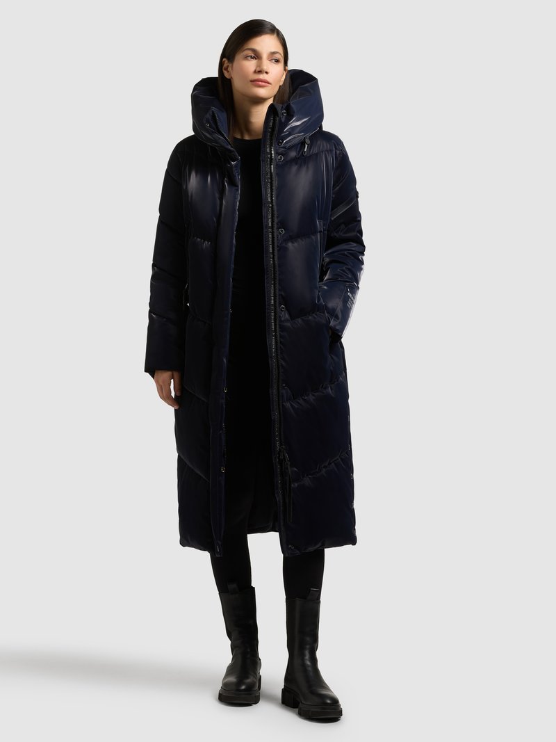 Dolga temno modra puhovka z visokim ovratnikom in kapuco, z lesketajočo se teksturo, diagonalnim šivom in stranskimi žepi; nošena preko črne obleke.