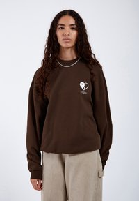 Felpa marrone con una vestibilità rilassata, maniche lunghe e una piccola grafica a forma di cuore bianco sul petto. Abbinata a pantaloni beige.