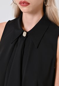 Primo piano del collo e della spalla di una donna che indossa una blusa nera, con una spilla decorativa al colletto e grandi orecchini a cerchio.