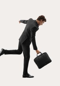 Schwarze Lederaktentasche mit zwei Griffen. Subjekt in einem maßgeschneiderten schwarzen Anzug, schreitet voran und zeigt glattes Material und präzise Schneiderei.