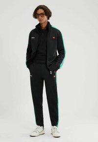 Chándal negro con rayas laterales verdes, chaqueta con cremallera, pantalones con cintura elástica, acentos de logo en la chaqueta y los pantalones, combinados con zapatillas blancas.
