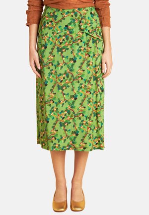 Grüner Midirock mit floralem und Astmuster, kombiniert mit einem rostfarbenen Langarmshirt und goldenen flachen Schuhen.