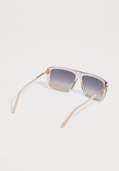 Lunettes de soleil carrées avec montures beige translucides et verres dégradés gris reposant sur une surface blanche.