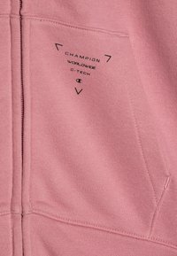 Sweatshirt rose avec une fermeture éclair et une poche avant, fabriqué en tissu doux. Les détails de marque notables incluent "Champion" et "c-tech" en noir.