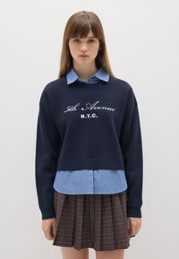 Marineblauer kurzer Pullover mit bestickter Aufschrift "5th Avenue N.Y.C.", über einem hellblauen gestreiften Hemd getragen, kombiniert mit einem braunen karrierten Faltenrock.
