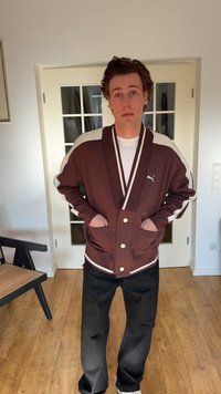 Mladý muž s kučeravými vlasmi, oblečený v hnedo-bielej bunde značky Puma a čiernych džínsach, stojí vo vnútri s rukami v vreckách bundy.