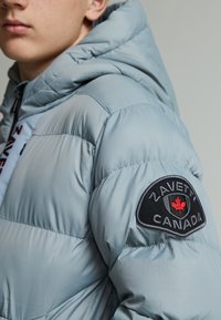Veste matelassée bleu clair avec capuche, présentant une texture matelassée, une fermeture éclair et un patch circulaire noir avec "ZAVETTI CANADA" et un motif de feuille.