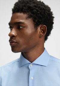 Vaaleansininen ruudullinen paita, jossa on jämäkkä kaulus, valkoiset napit ja sileä tekstuuri, päällä henkilöllä, jolla on lyhyet, kiharat hiukset.