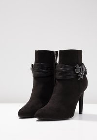 Botines de ante negros con punta afilada, tacón de aguja y detalle decorativo de hebilla en el lado con una correa suave. Cierre de cremallera.