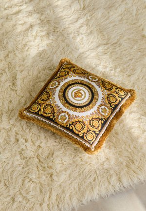 Decorative pillow with a gold and black patterned design, featuring a soft, fringed edge, placed on a fluffy white rug.