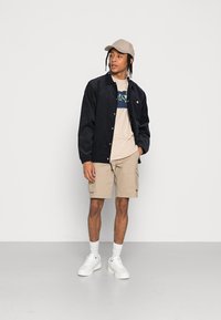 Schwarze Cordjacke, beiges T-Shirt mit Grafik, beige Cargo-Shorts und weiße Sneakers. Das Modell trägt eine hellbraune Kappe mit Zöpfen.