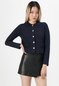Vrouw met lang steil haar, gekleed in een marineblauw vest met knopen en een zwarte leren minirok, staand met één hand op de heup.