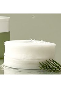 Witte, ronde wattenpad op een glanzend oppervlak, met een subtiele textuur. Een groene container en een takje groen zijn zichtbaar op de achtergrond.