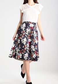Falda plisada floral en azul marino oscuro con flores rojas, blancas y verdes. Tela ligera, diseño en capas, combinada con una blusa blanca y zapatos negros.