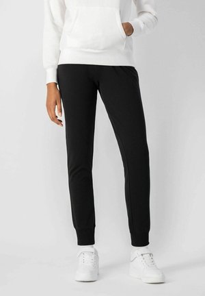 Pantalones de chándal negros ajustados, hechos de una tela suave, con una textura lisa, cinturilla elástica y puños acanalados, combinados con deportivas blancas.