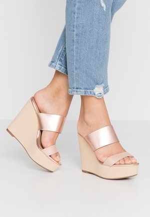 Sandały na koturnie z błyszczącego różowego złota z szerokim paskiem oraz beżową platformą. Otwarte palce, gładka faktura, zestawione z przetartymi niebieskimi dżinsami.