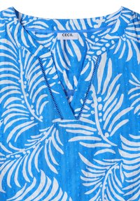 Chemise bleue avec motif de feuilles tropicales blanches, col en V avec bordure cousue, et étiquette de marque "CECIL" au col.