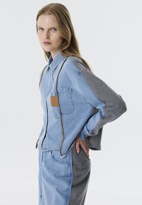 Jeansjack met grijze mouwen en een blauwe romp, voorzien van een borstzak, gestikt detail en een contrasterende bruine patch op de zak.