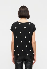 Άτομο με κοντά σκούρα μαλλιά που φοράει μαύρο μπλουζάκι με λευκά σχέδια καρδιών και μαύρο δερμάτινο παντελόνι, φαίνεται από πίσω.