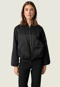 Černá bomber bunda s hladkou, lesklou texturou. Má zipový přední díl, žebrovaný límec a elastický lem. Dlouhé, nadýchané rukávy.