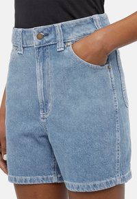 Calções de ganga azul claro com cintura alta, apresentando bolsos frontais, fecho de botão metálico e detalhes de costura branca. Textura suave.