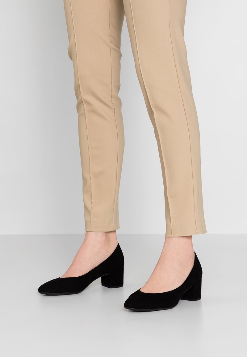 Chaussures à talons carrés en daim noir avec un design à bout rond, associées à un pantalon beige ajusté avec des détails de couture visibles.