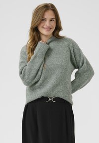 Jonge vrouw met lang haar in een gezellige grijze gebreide trui en zwarte broek, glimlachend en poserend met één hand bij haar gezicht.