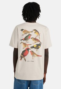 Μπεζ μπλουζάκι με κοντά μανίκια που διαθέτει διάφορες πολύχρωμες εικόνες πουλιών τυπωμένες στην πλάτη, παρουσιάζοντας διαφορετικά σχήματα και σχέδια.