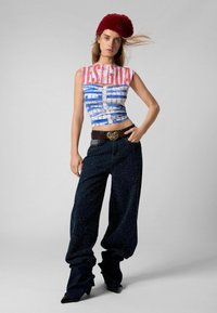 Grafikus tank top kék és fehér csíkokkal, rajta a "VESTIGUA" felirattal. Laza sötét farmernadrággal és bordó prémkalapral párosítva.