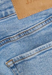 Tecido de denim azul claro com textura e costura visíveis, apresentando um emblema de couro castanho e dois bolsos frontais.