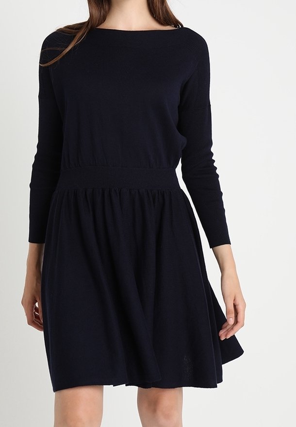 Woman wearing a long-sleeve, knee-length, dark navy blue dress with a fitted waistband and slightly flared skirt.