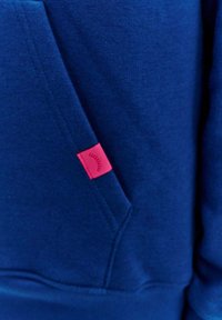 Tmavě modrá mikina vyrobená ze směsového bavlněného materiálu. Má přední kapsu a malou růžovou visačku s grafickým detailem. Hladký povrch.