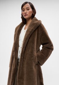 Casaco de pêlo falso castanho com um colarinho largo, bolsos frontais e um design aberto, sobre um top branco com detalhes de rendas.