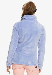 Vaaleansininen fleece-takki, jossa on korkea kaulus, pitkät hihat ja pehmeä pinta. Takissa on sivutagi helmassa. Takakatsaus vaatteeseen.