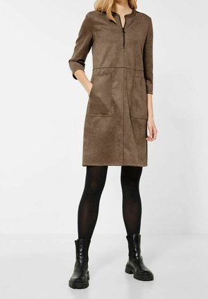 Žena v hnědých semišových šatech po kolena s předními kapsami, tříčtvrtečními rukávy, černými punčochami a černými robustními botami, stojící na bílém pozadí.