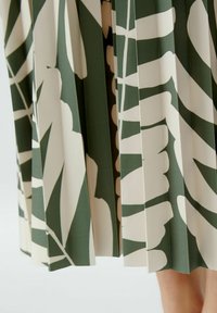 Jupe plissée verte et crème avec un motif de feuilles, fabriquée dans un tissu à la texture lisse. Présente des designs géométriques nets et de profonds plis.