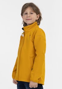 Gul softshell-jakke med høj krave, lynlåslukning, to sidelommer og dekorative lynlåse, med et logo på brystet.