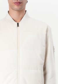 Helle beige gesteppte Jacke mit Reißverschluss, geripptem Kragen und glatter Oberfläche. Präsentiert ein minimalistisches Design und ein kleines Logo am Ärmel.