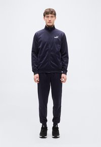 Σκούρο μπλε φόρμα με ζακέτα με φερμουάρ και παντελόνι με ελαστική μέση. Διαθέτει λογότυπο στο στήθος και αθλητικά παπούτσια. Ομαλό, ελαφρύ υλικό.